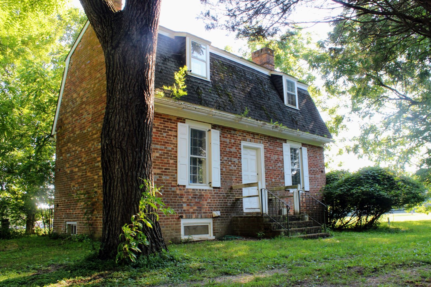 Looking up from the back-left lawn beneath the shade of a mature tree. This low angle emphasizes the sturdy, compact footprint of the two-story colonial homestead and the steep slope of its roof.